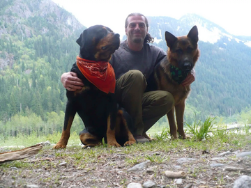Douglas Spink photographed outdoors, crouching with both arms resting on a dog at each side